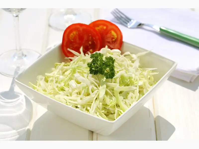 Frischer, selbstgemachter Krautsalat in einer weißen Schüssel, garniert mit Petersilie und Tomatenscheiben. Daneben liegen ein grüner Löffel und eine Serviette.