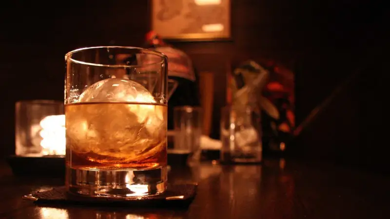 Ein Glas mit bernsteinfarbenem Whisky und einer großen Eiskugel steht auf einem dunklen Holztisch. Man trinkt Whisky mit Eis für den perfekten Genuss.