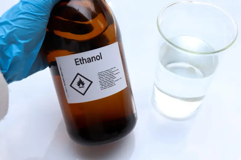 Braune Glasflasche mit der Aufschrift "Ethanol" und einem Warnsymbol. Daneben ein Becherglas mit klarer Flüssigkeit. Hier gibt es 100 prozentigen Alkohol.